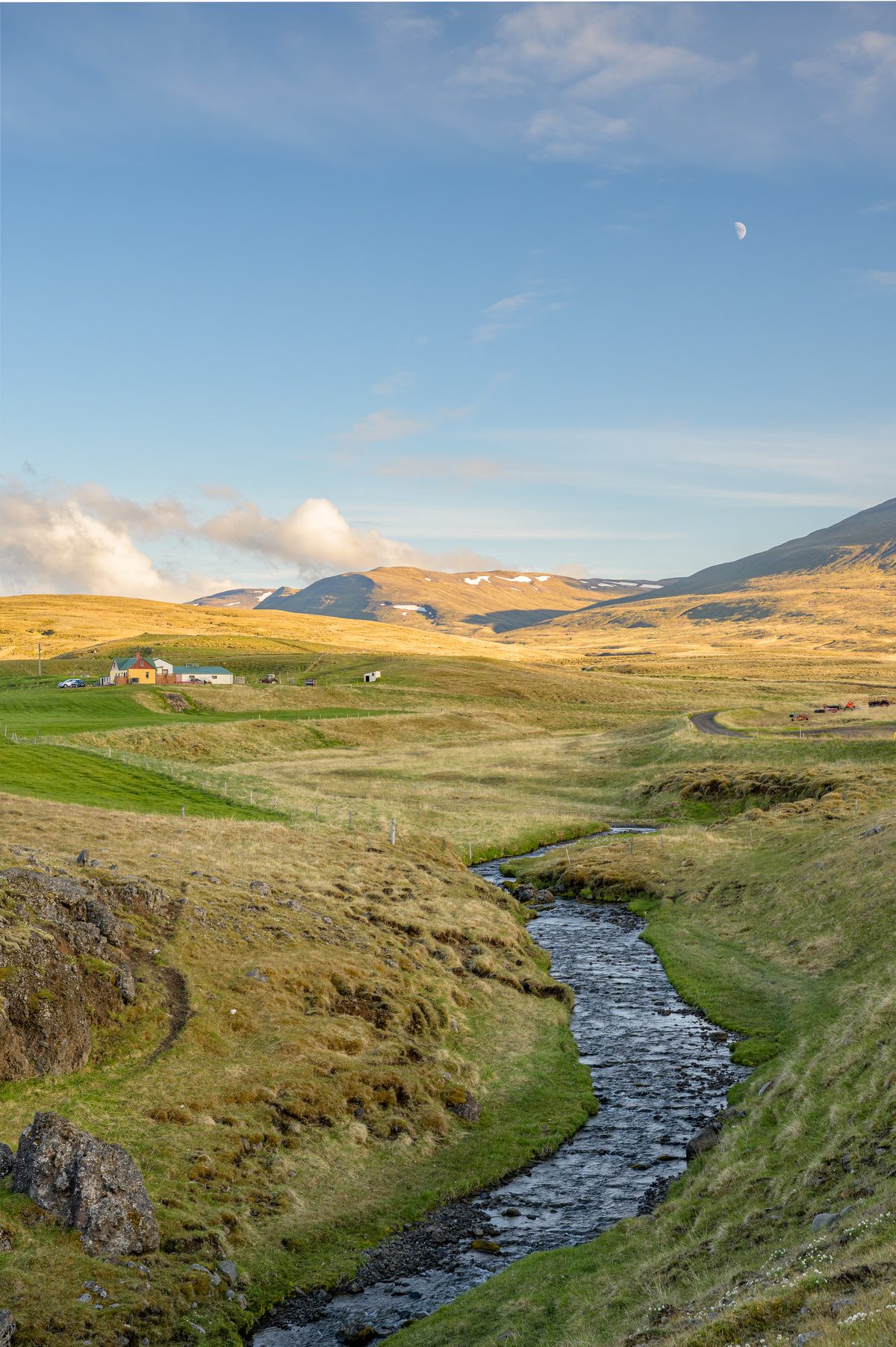 A narrow stream winds through a grassy volcanic hollow toward sunlit hills beneath a pale blue sky.