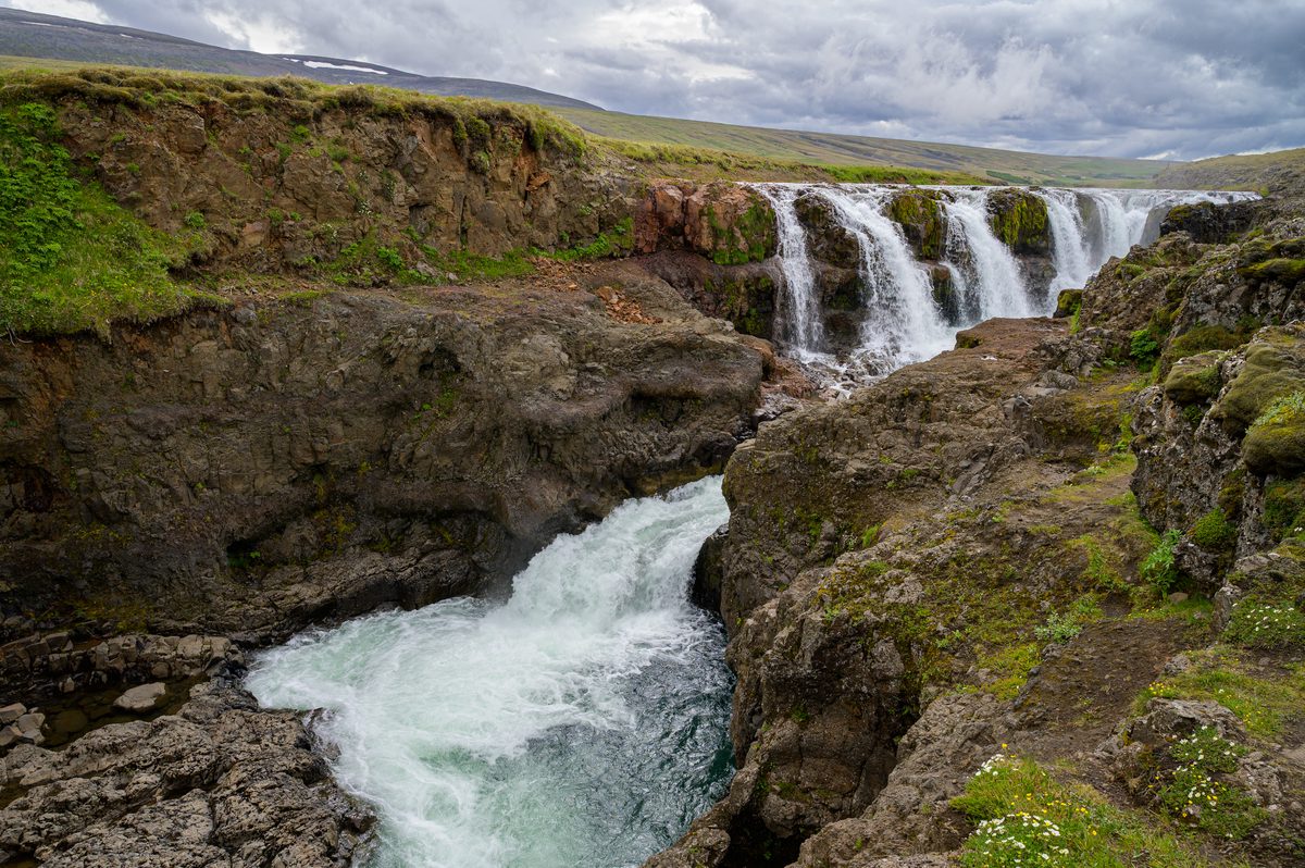 Several curtains of white water spill over a rocky ledge at Kolufossar before rushing through the narrow gorge beneath a heavy gray sky.
