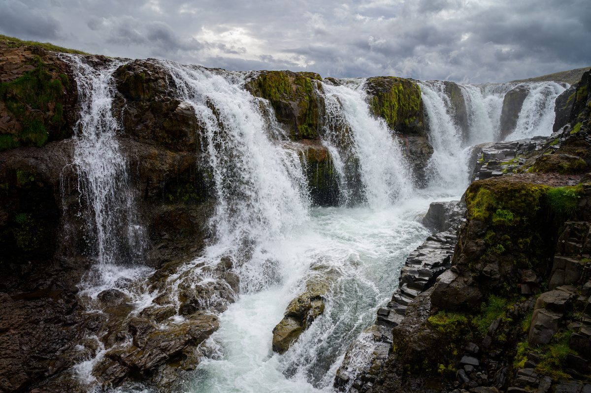 A broad, close view of Kolufossar captures the falls dividing around dark, moss-covered rocks and churning through the channel below.