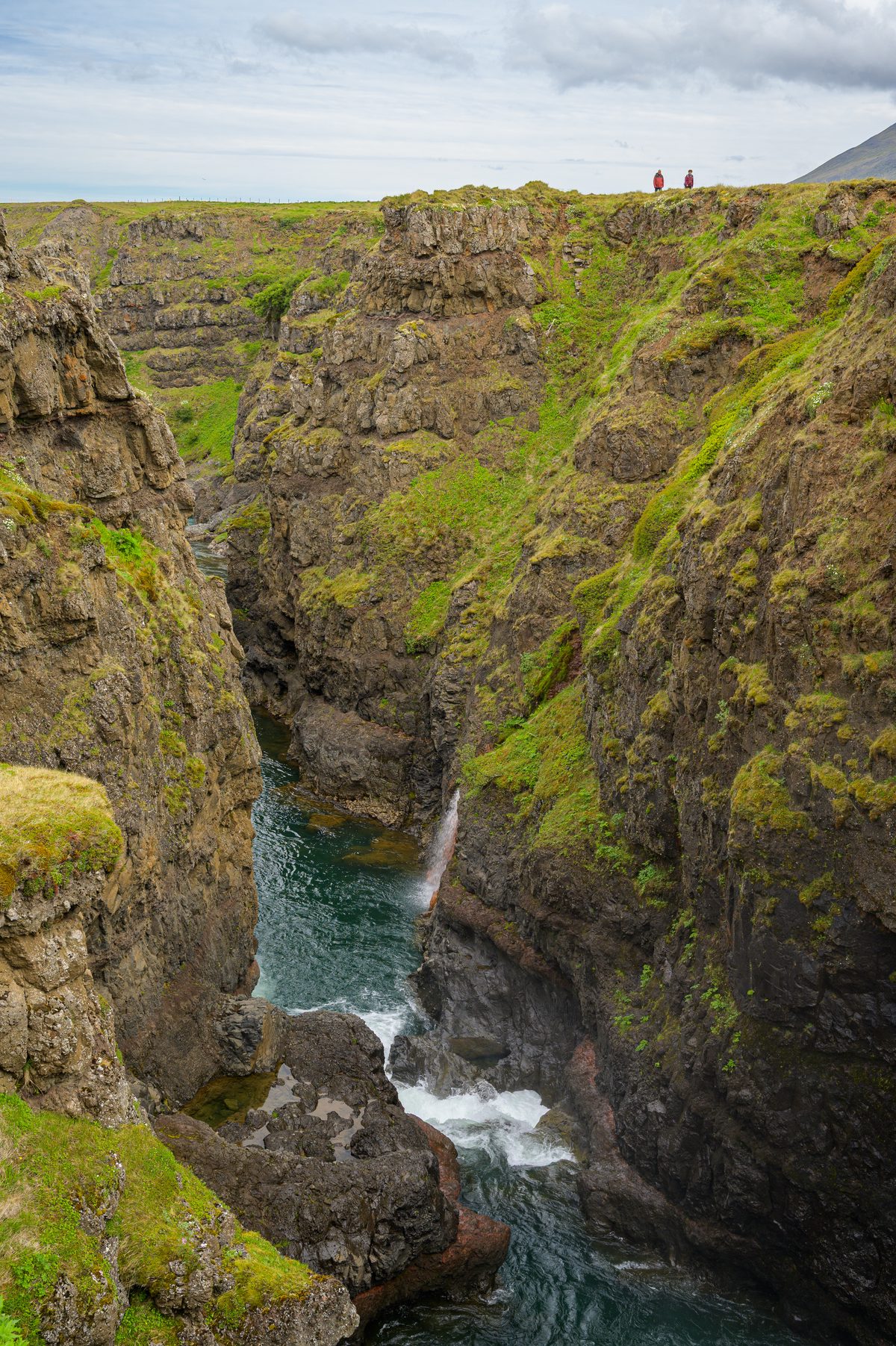 A vertical view looks down the steep, green-streaked walls of Kolugljúfur to a turquoise river and small cascades, with tiny visitors standing on the far rim.