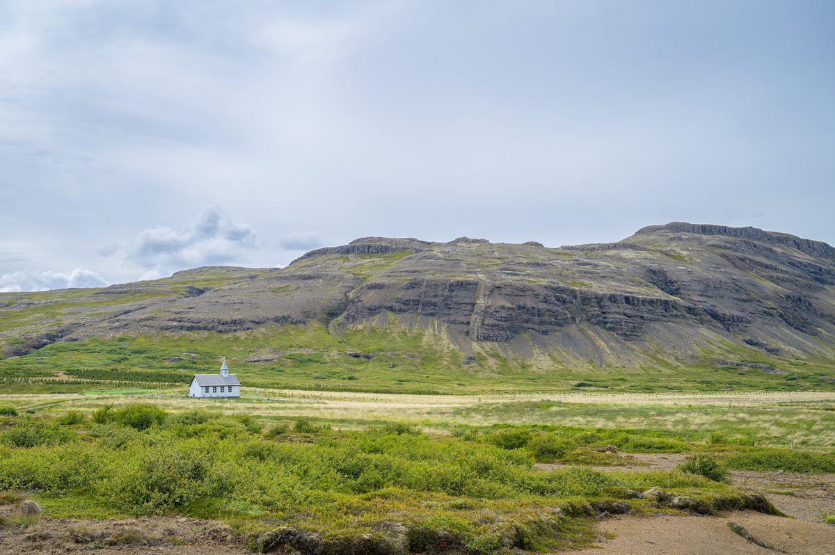 A small white church stands in pale green fields below a broad mountain patterned with horizontal bands of dark rock.