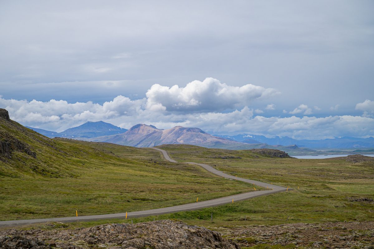 An empty paved road curves across open green and brown moorland toward distant mountains lit beneath a bank of billowing clouds.