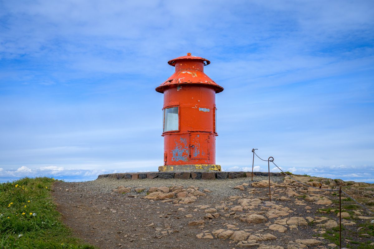 The weathered orange lighthouse on Súgandisey rises from a rocky platform beneath a blue sky brushed with thin clouds.