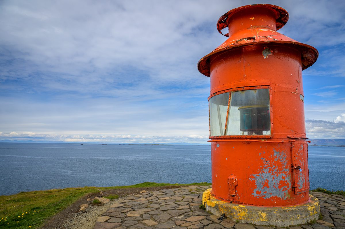A close view of Súgandisey lighthouse reveals peeling orange paint and its curved lantern window, with blue water stretching to the horizon.
