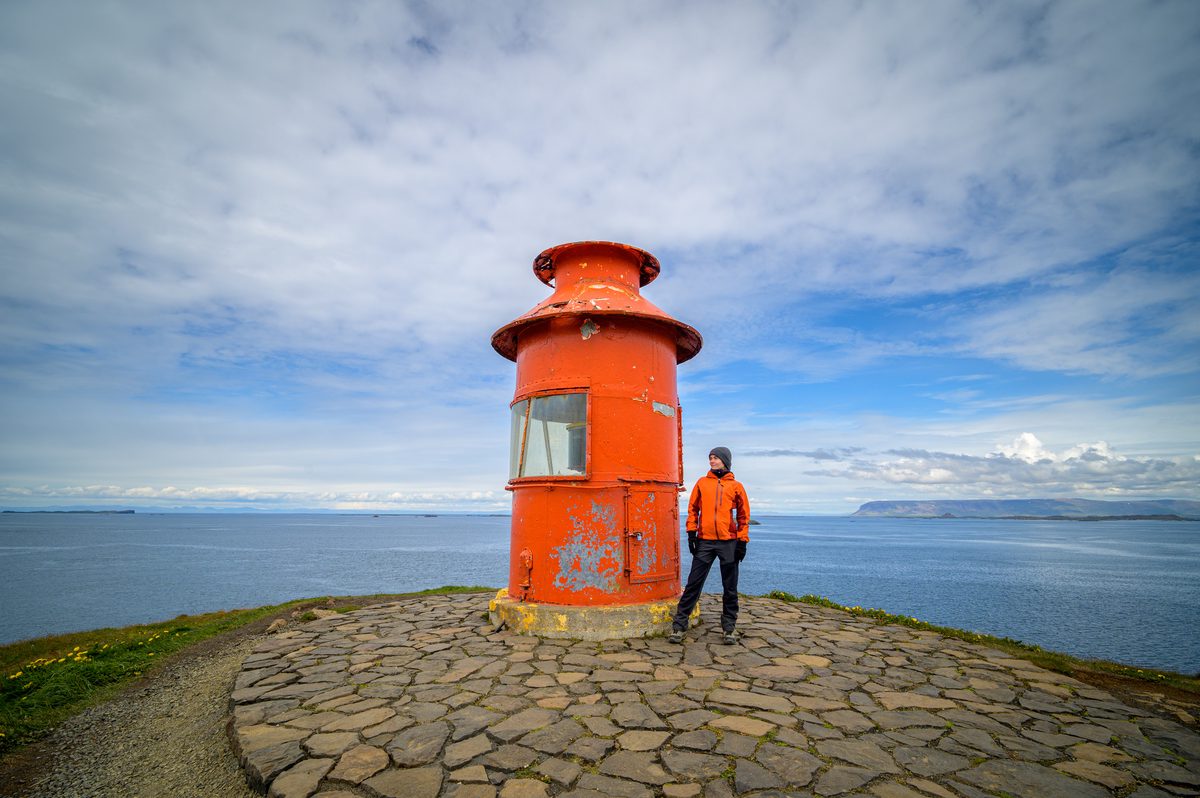 A traveler wearing an orange jacket stands beside the orange Súgandisey lighthouse on a stone-paved lookout above the sea.