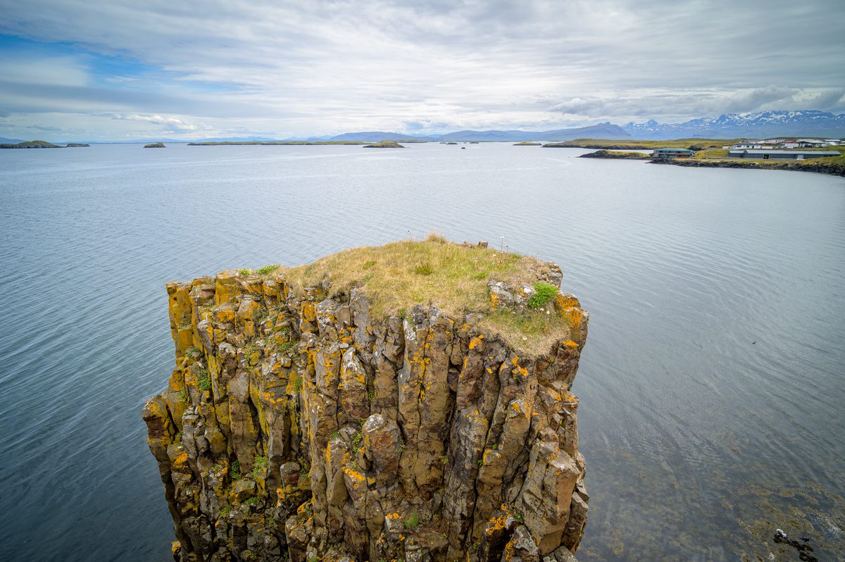 A grass-topped outcrop of vertical basalt columns rises above still water, with scattered islands, distant buildings, and snow-streaked mountains beyond.