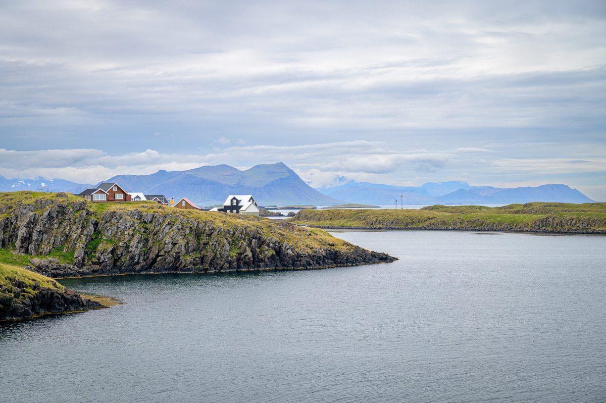 A cluster of houses sits on low, dark coastal cliffs across a sheltered inlet, backed by distant blue-gray mountains.