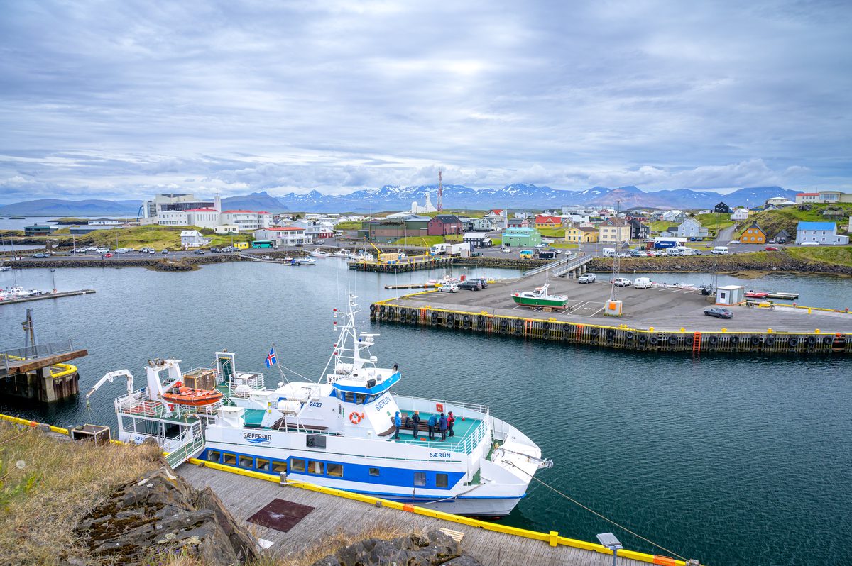 A white passenger boat with blue and yellow trim is moored below a view across Stykkishólmur harbour, its colorful buildings, quays, and distant mountains.