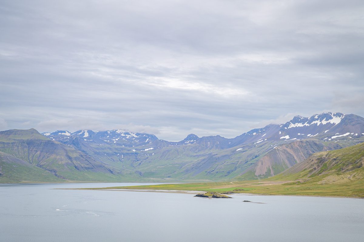 Calm water fills the foreground beneath a sweeping mountain ridge, where lingering snow marks gullies above green lower slopes.