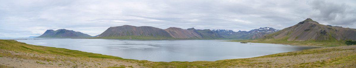 A wide panorama follows a quiet inlet around a chain of rugged mountains, with grassy shoreline in the foreground and layered gray clouds overhead.