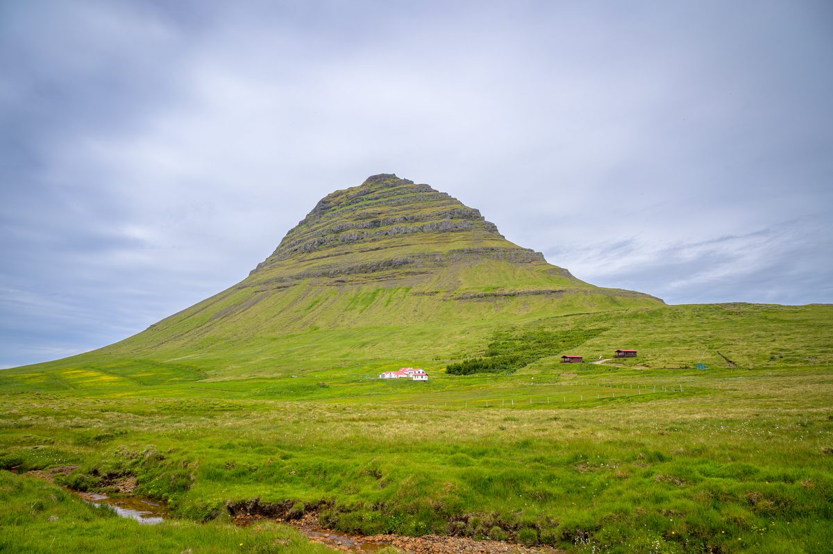 Kirkjufell fills the view with its stepped bands of rock and bright green slopes, while small farm buildings sit near the foot of the mountain.