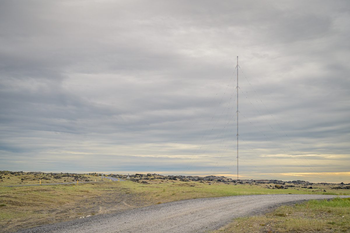 A very tall, slender radio mast rises above a gravel track and low lava-strewn ground, with a narrow strip of warm light beneath heavy clouds.