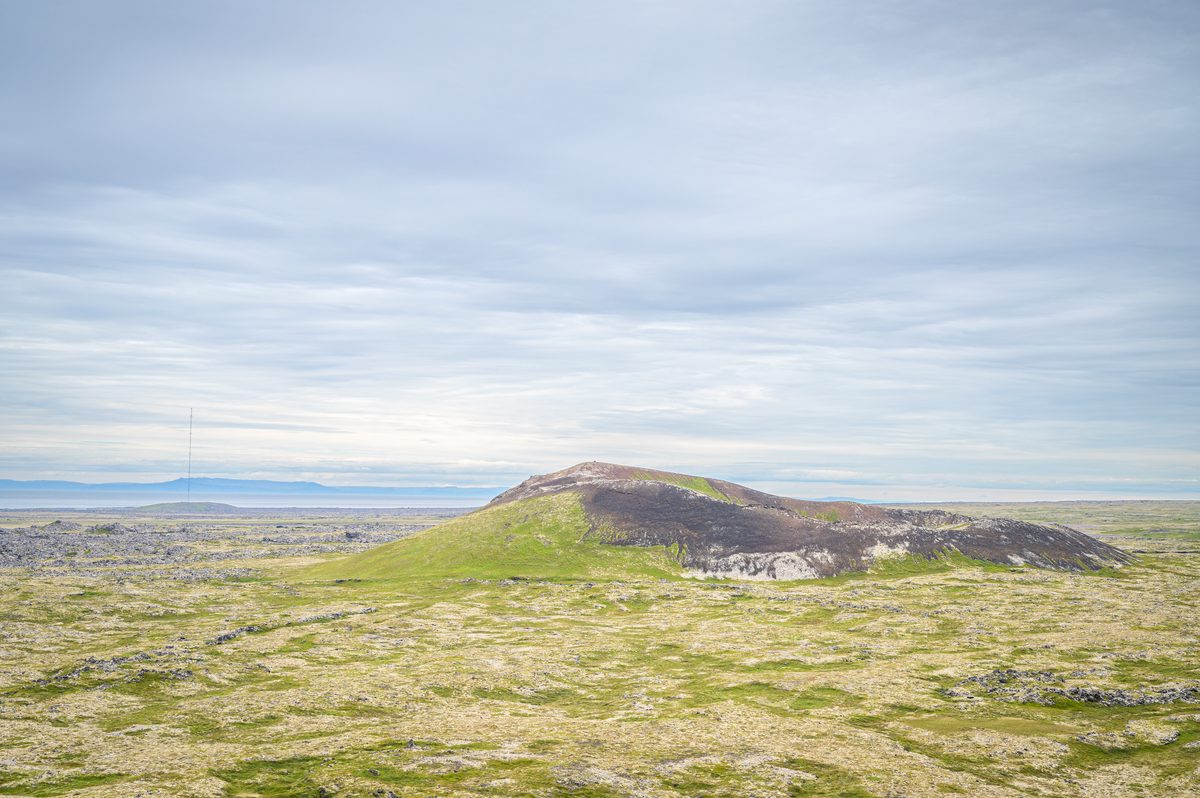 A low volcanic mound streaked with green vegetation and pale rock stands amid a broad lava plain beneath a softly layered sky.