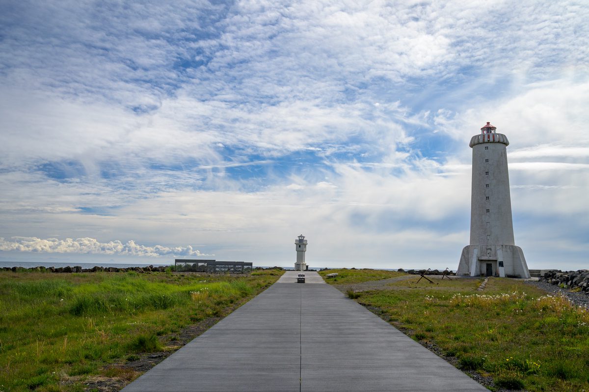 A straight paved path crosses green grass toward the small old lighthouse at Akranes, while the taller white lighthouse rises to the right beneath a textured sky.