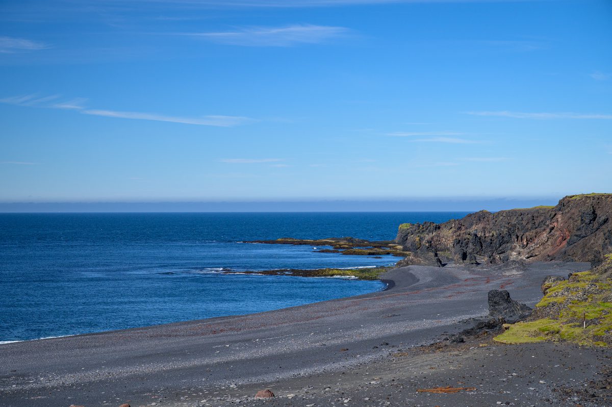 A dark volcanic beach curves beneath rugged coastal cliffs beside a deep blue sea, with scattered rocks and green patches along the shore.