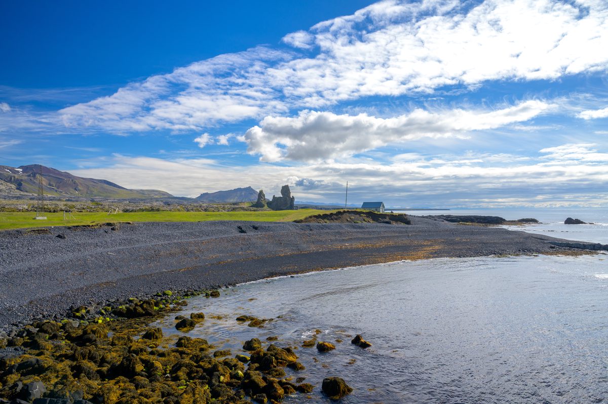Still water borders a dark pebble beach at Arnarstapi, with seaweed-covered rocks in the foreground and jagged coastal formations beneath the mountains beyond.