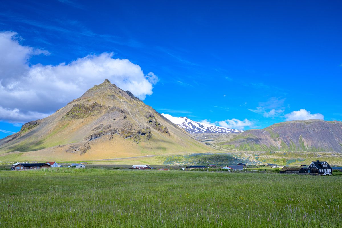 The pointed, layered slopes of Stapafell rise above green grass and scattered buildings at Arnarstapi, with snow-covered peaks visible behind.