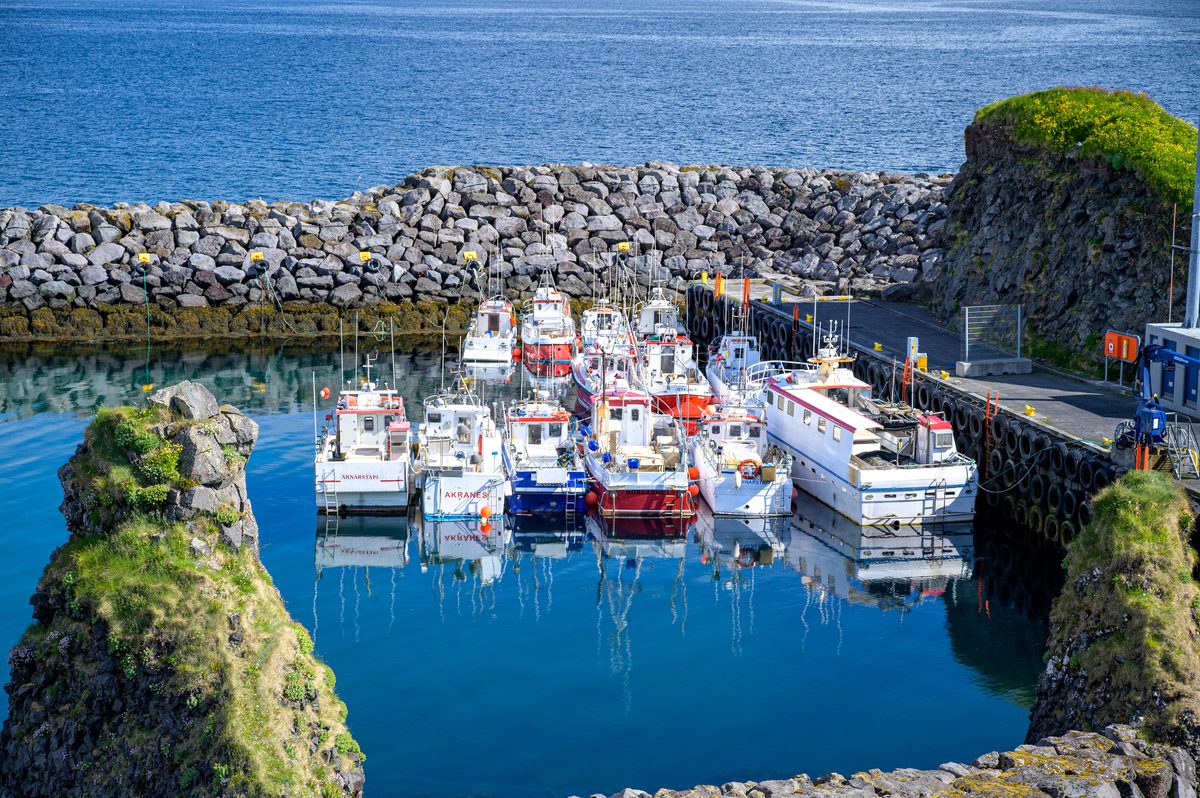 Small fishing boats cluster in the still blue water of Arnarstapi harbour, enclosed by steep rocky sides and a pale stone breakwater.