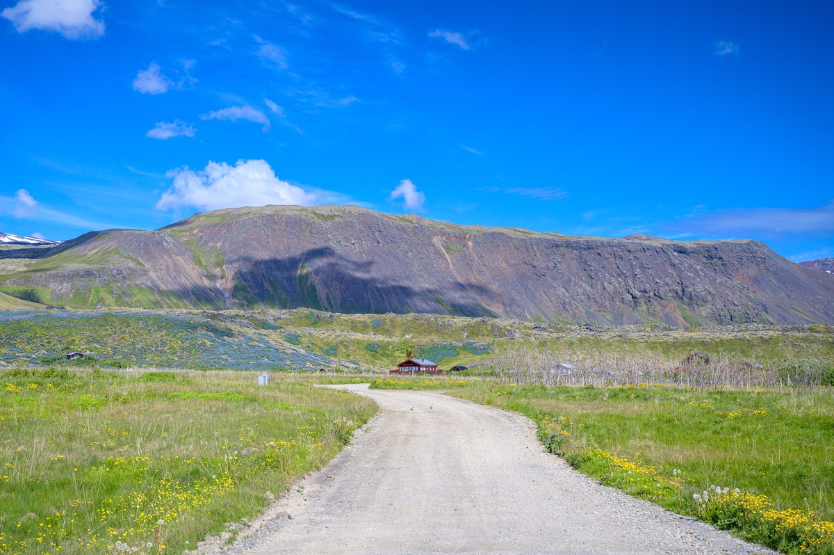 A pale gravel road curves through green grass and low vegetation toward scattered buildings beneath a broad, steep-sided mountain ridge.