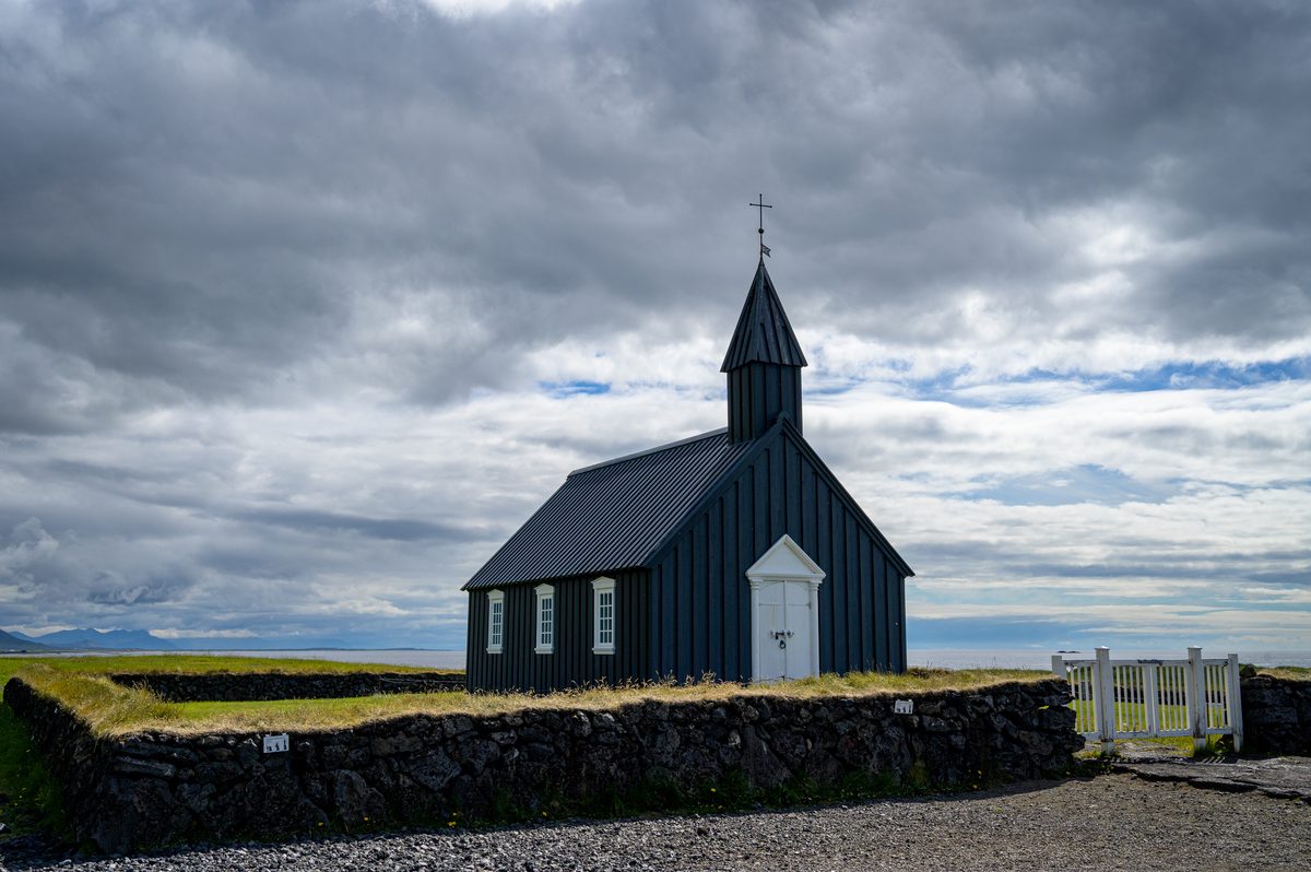 The black wooden church at Búðir stands behind a low stone wall, its white door and windows contrasting with the dark boards and gray clouds.