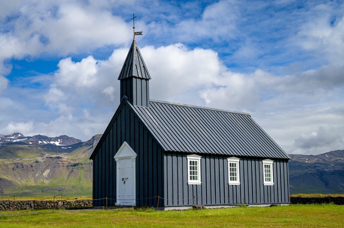 A close three-quarter view of Búðakirkja shows its white entrance, narrow windows, and dark steeple against a mountain backdrop and broken clouds.