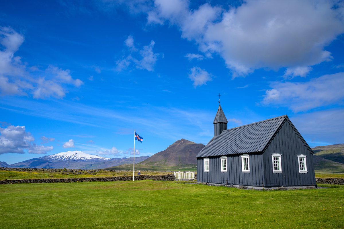 Búðakirkja stands across a wide green lawn, with an Icelandic flag, a low stone wall, and snow-covered mountains beneath a bright blue sky.