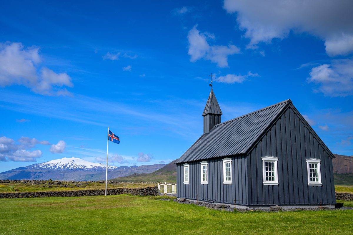 The side and rear of Búðakirkja reveal rows of white-framed windows above a sunlit lawn, with an Icelandic flag and snowy peaks to the left.
