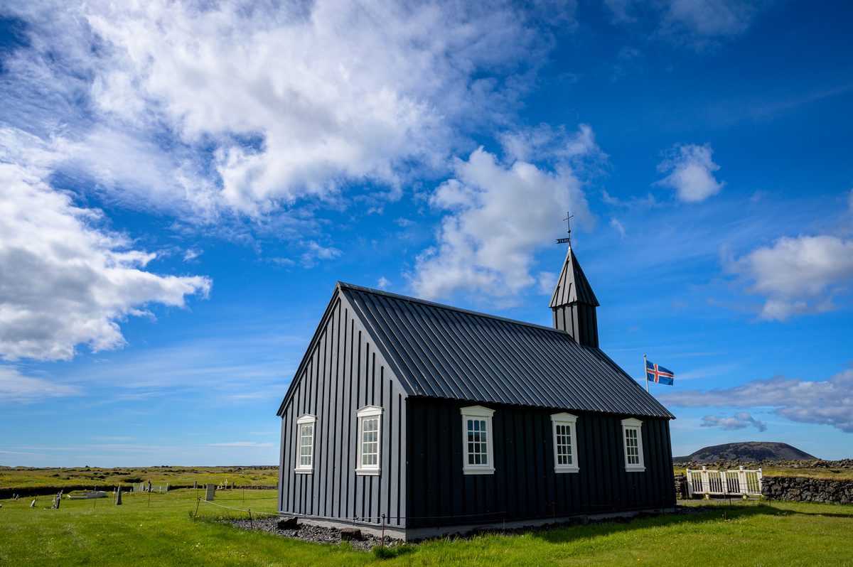 A rear three-quarter view of Búðakirkja sets its black timber walls and white windows beneath a broad sky of blue and white, with gravestones visible beside the church.