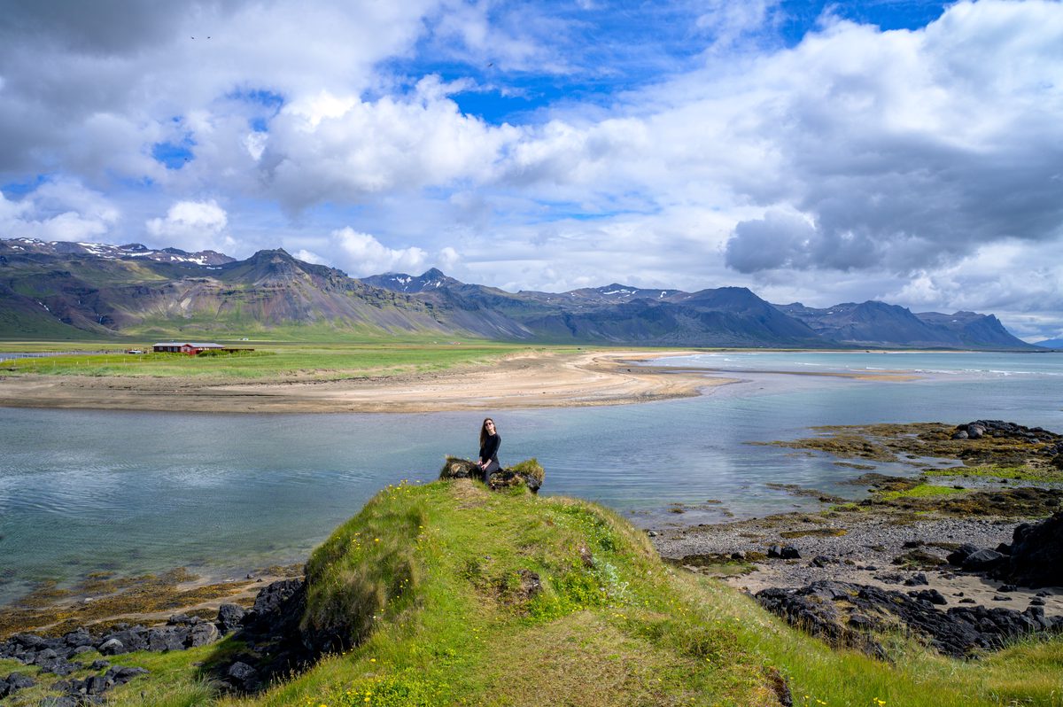 A person stands on a small grassy outcrop above a calm bay, looking toward a pale sandy shoreline and a long ridge of rugged mountains.