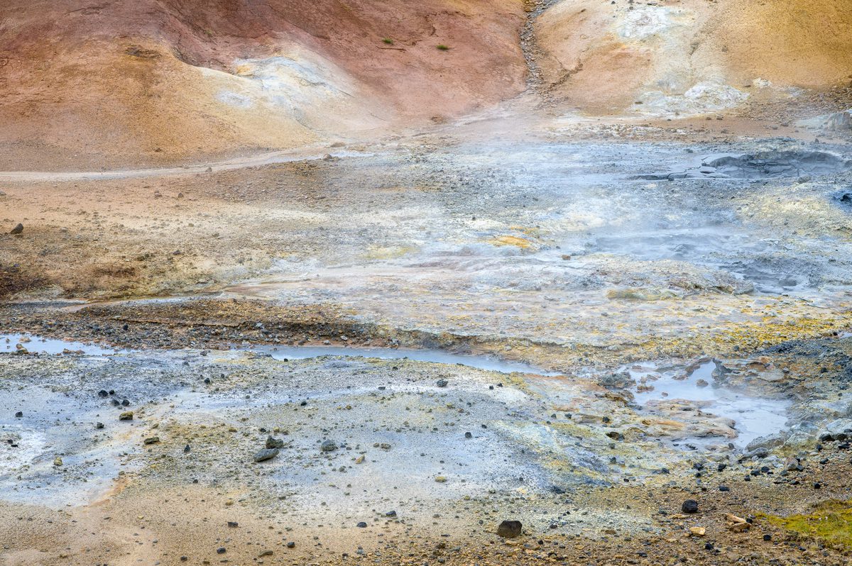 Wisps of steam drift across shallow pools and mineral-stained ground at Seltún, beneath slopes streaked with ochre, rust red, and pale yellow.