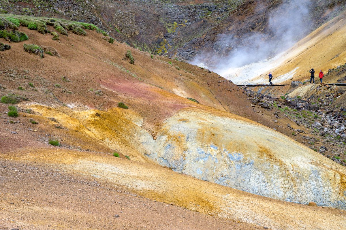 Visitors follow a boardwalk beside steaming vents at Seltún, beyond rounded slopes patterned with yellow, white, and reddish-brown earth.