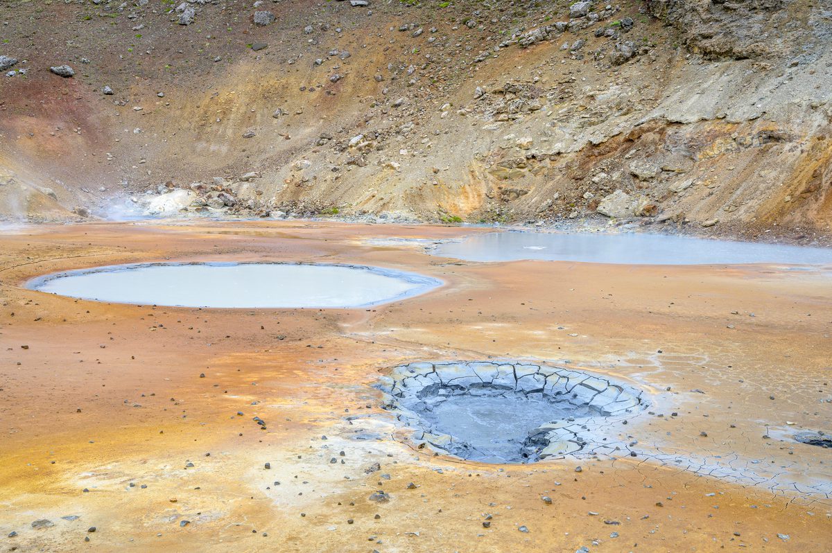 Pale blue-gray pools interrupt the orange geothermal ground, with cracked mud around a small foreground vent and steam rising at the foot of the rocky slope.