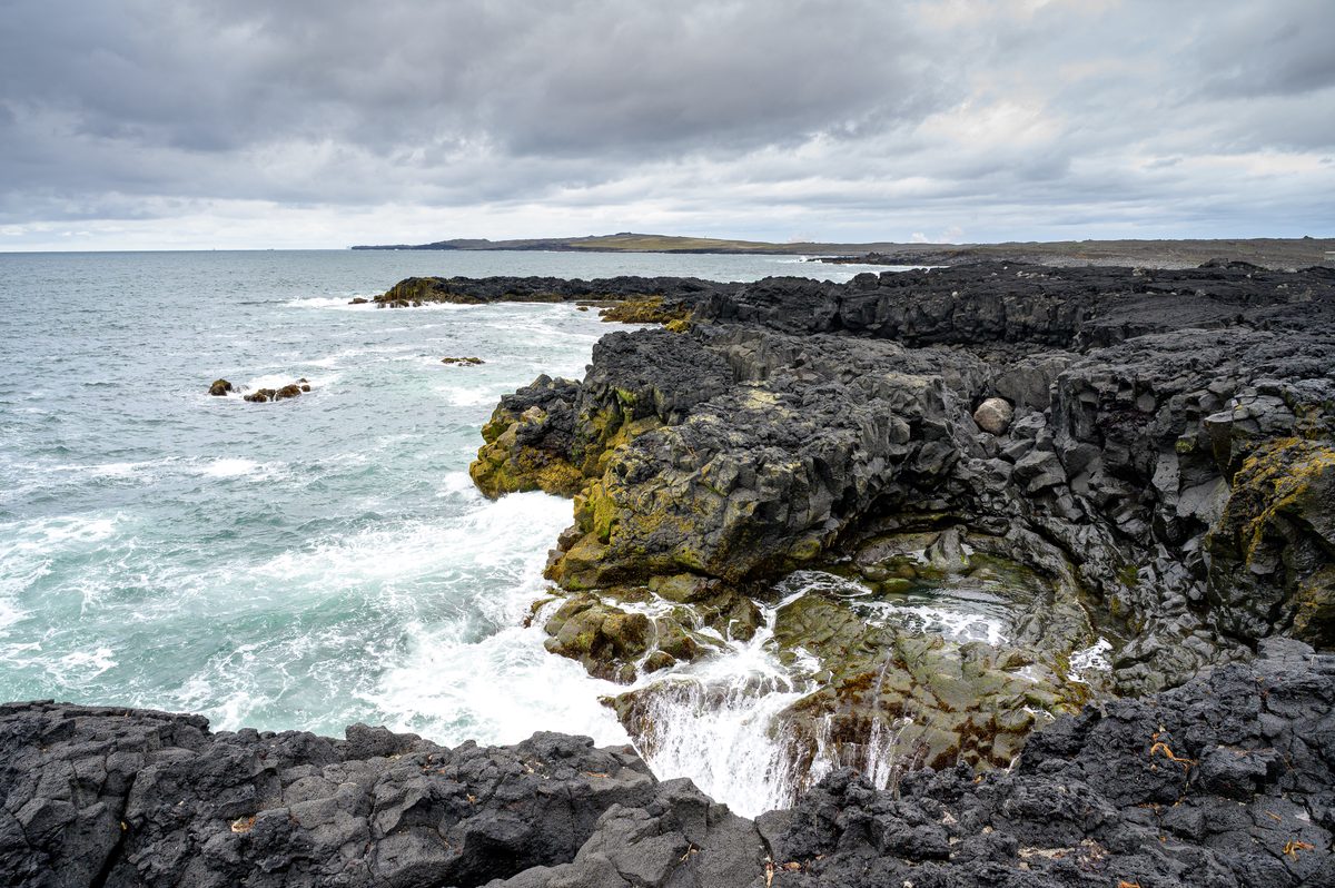 White surf washes into a narrow inlet in the jagged black lava coast, where yellow-green seaweed marks the rocks beneath a heavy gray sky.