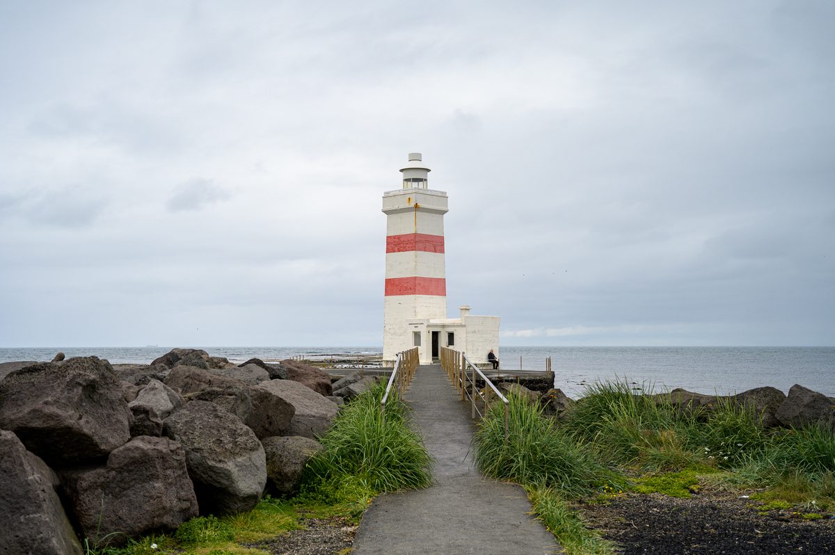 A narrow path and wooden footbridge lead between grass and dark boulders to the old white lighthouse at Garðskagi, its two faded red bands standing out against the gray sea and sky.